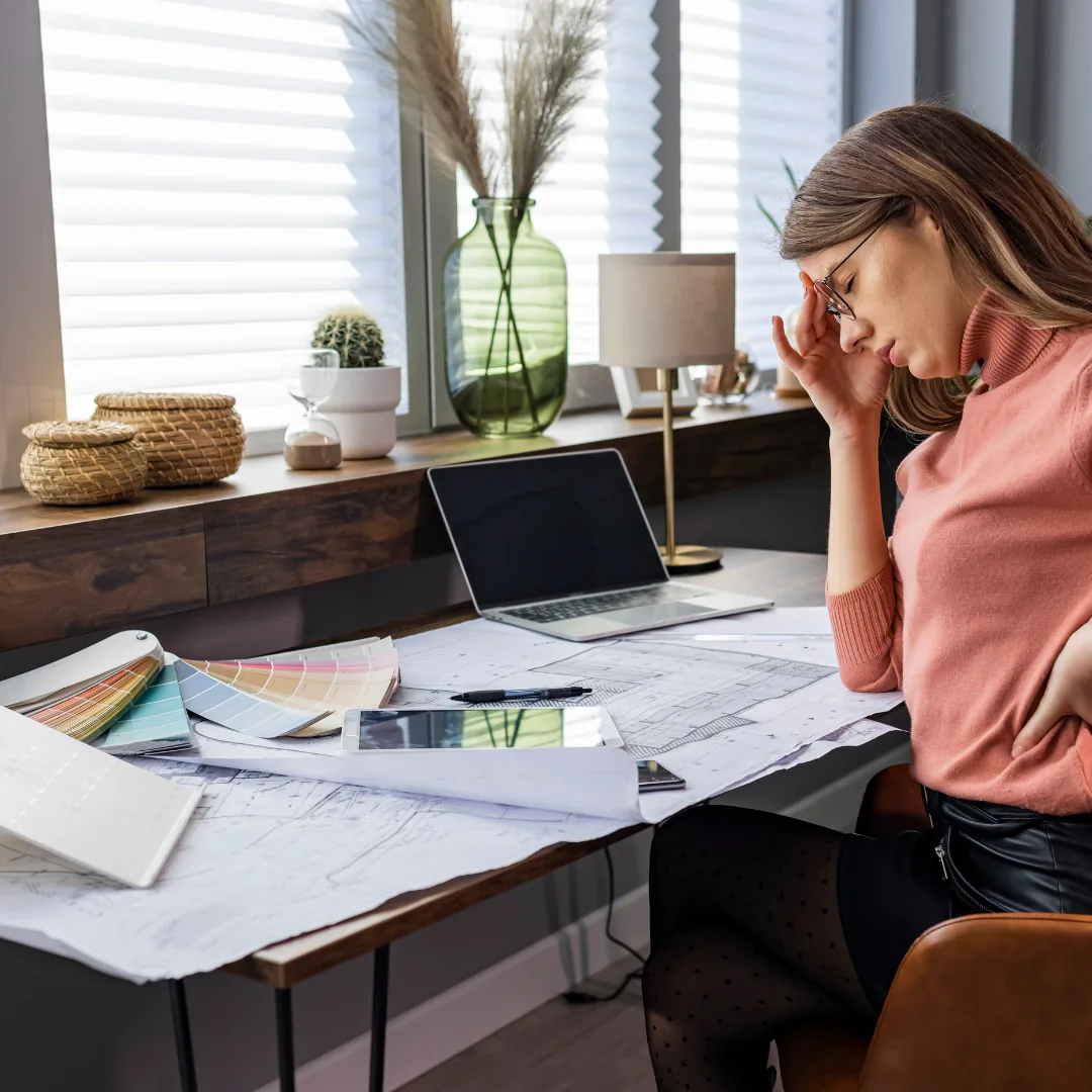 Woman at a desk holding her temple while leaning over large paper plans, a laptop, tablet, and color swatches.