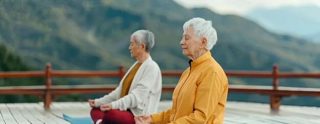 Two older adults seated cross-legged on yoga mats meditating on a wooden deck with mountain views.