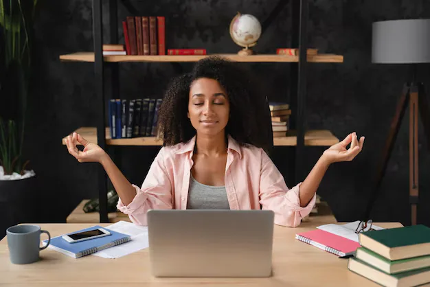 Woman seated at a desk with laptop and books practicing a calm meditation pose, eyes closed and hands in a mudra, suggesting brain-health self-care.