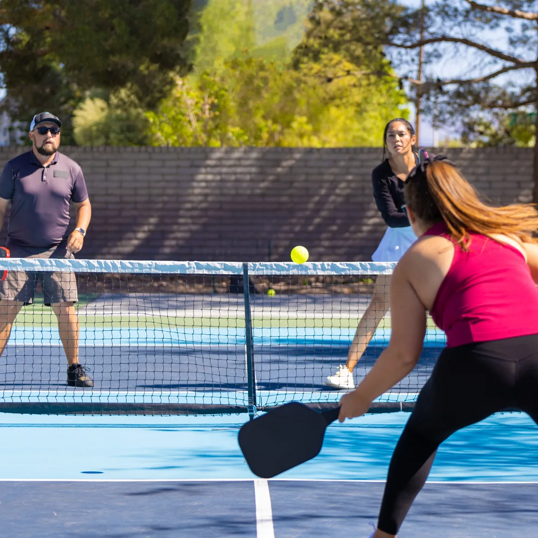 Two players returning a pickleball over a net on an outdoor court, paddle contacting the ball, suggesting light activity related to chronic fatigue and energy management.