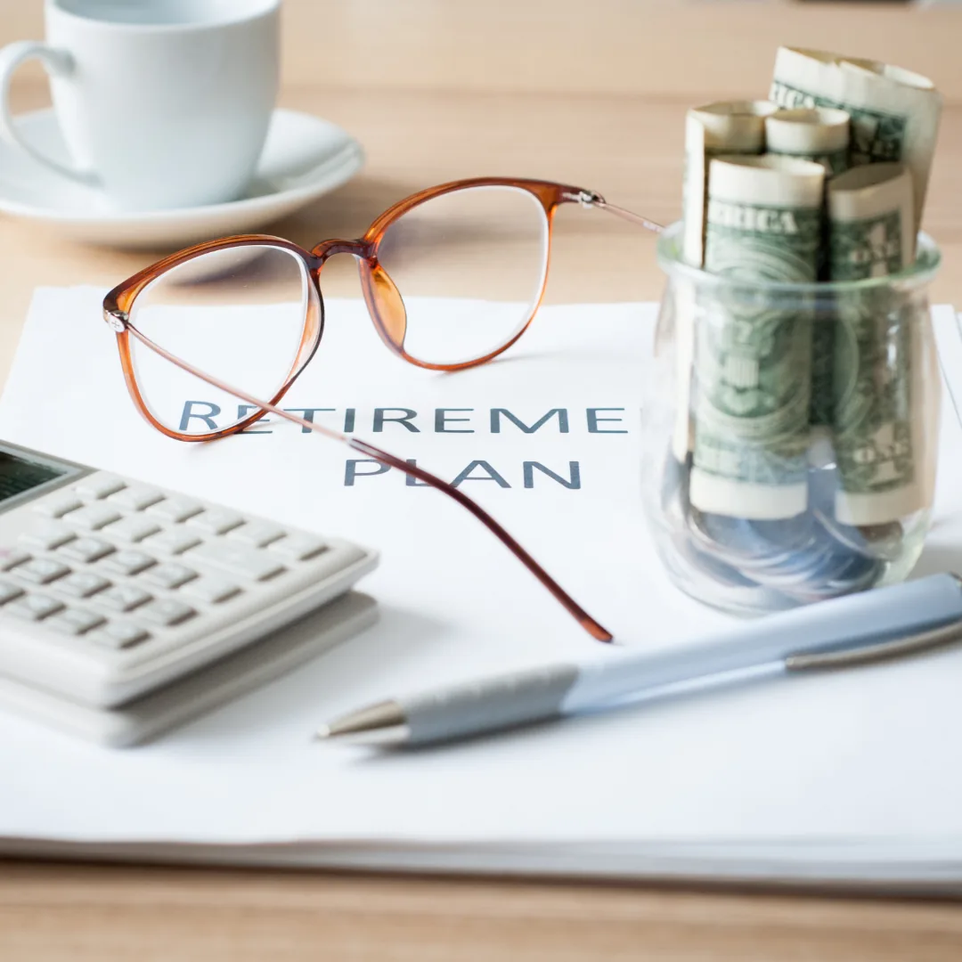 Reading glasses on a retirement plan document beside a calculator, pen, jar of rolled dollar bills and a coffee cup