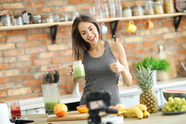 Person recording a gut-friendly green smoothie tutorial, holding a jar and giving a thumbs-up with fresh fruit, blender, and camera on the kitchen counter.
