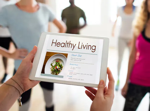 Hands holding a tablet displaying a 'Healthy Living' diet plan with a salad photo; blurred group exercising in the background.