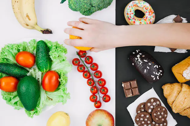 Hand reaching for an orange on a split surface showing fresh vegetables and fruit on the left and pastries, cookies, and chocolate on the right.