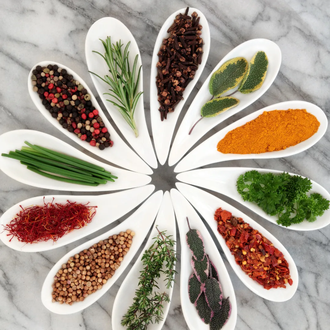 Assorted fresh and dried herbs and spices displayed in white ceramic spoons arranged in a circular pattern on a marble surface.