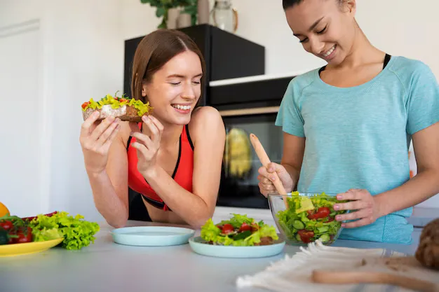 Two women preparing a fresh vegetable salad and serving vegetable-topped toast on a kitchen counter.