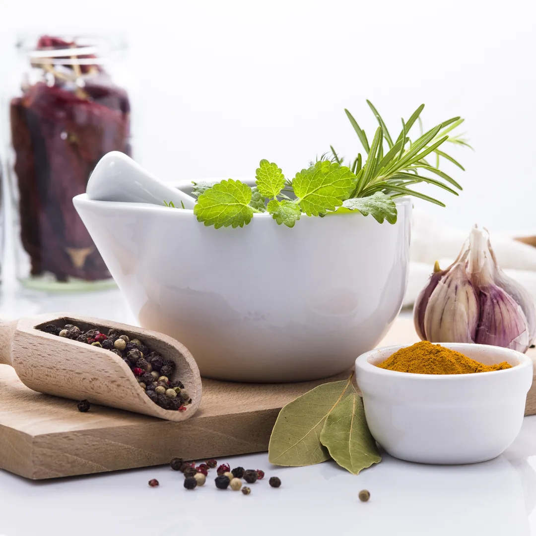 Ceramic mortar and pestle holding fresh herbs beside a bowl of turmeric, garlic, bay leaves, and a scoop of mixed peppercorns on a cutting board