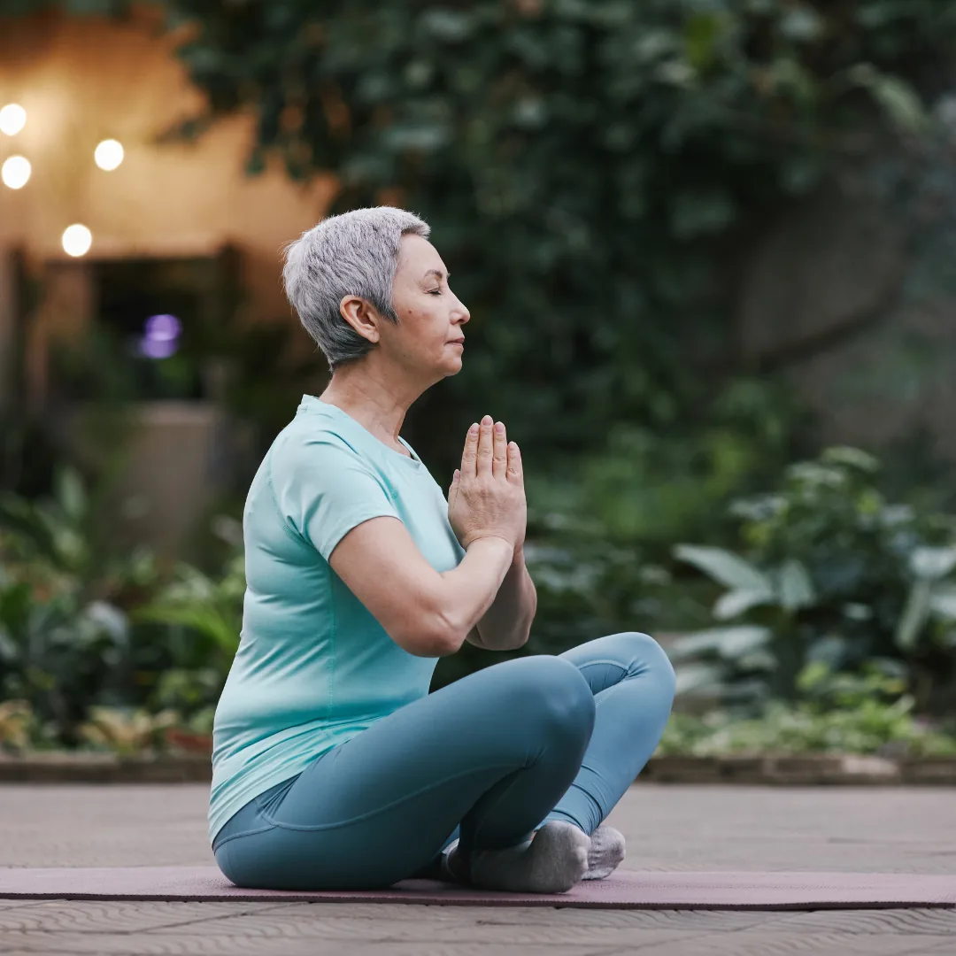 Person seated cross-legged on a mat outdoors with hands in prayer, meditating for stress management and longevity.