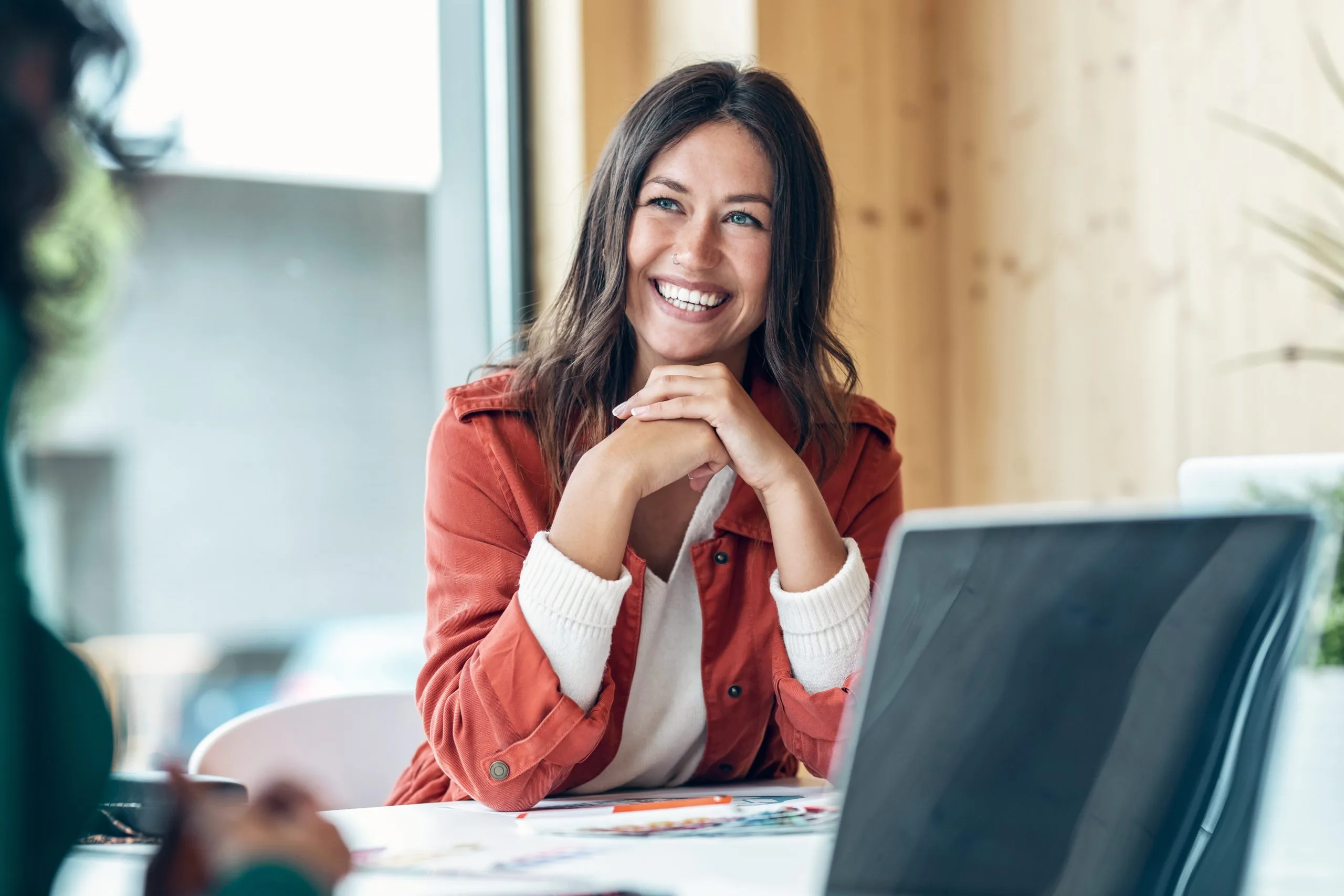 Smiling woman resting her chin on clasped hands at a desk with a laptop, engaged in conversation in a bright indoor workspace.