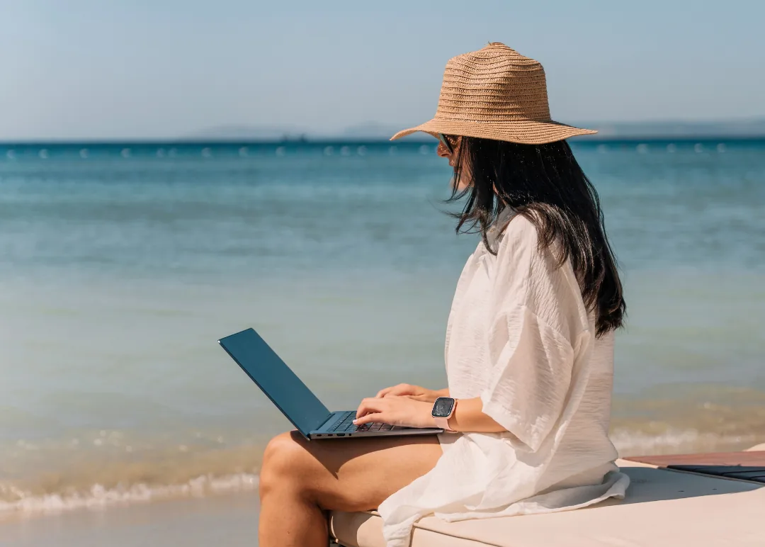Woman wearing a straw sun hat seated on a beach edge using a laptop with calm sea and horizon behind her