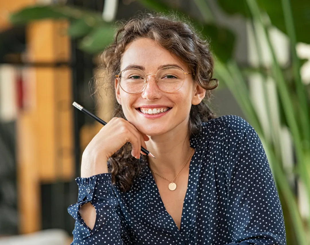 Smiling woman wearing glasses, holding a pencil and resting her chin on her hand in a relaxed indoor setting