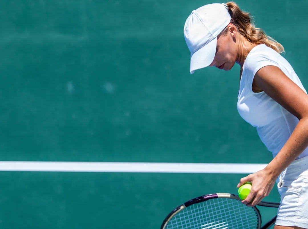 Tennis player leaning forward holding a ball and racket, preparing to serve on a green court.