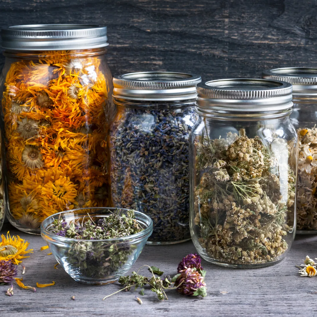 Glass jars and a small bowl holding assorted dried herbs and flowers on a wooden surface, displayed as natural remedies used in functional medicine for thyroid support