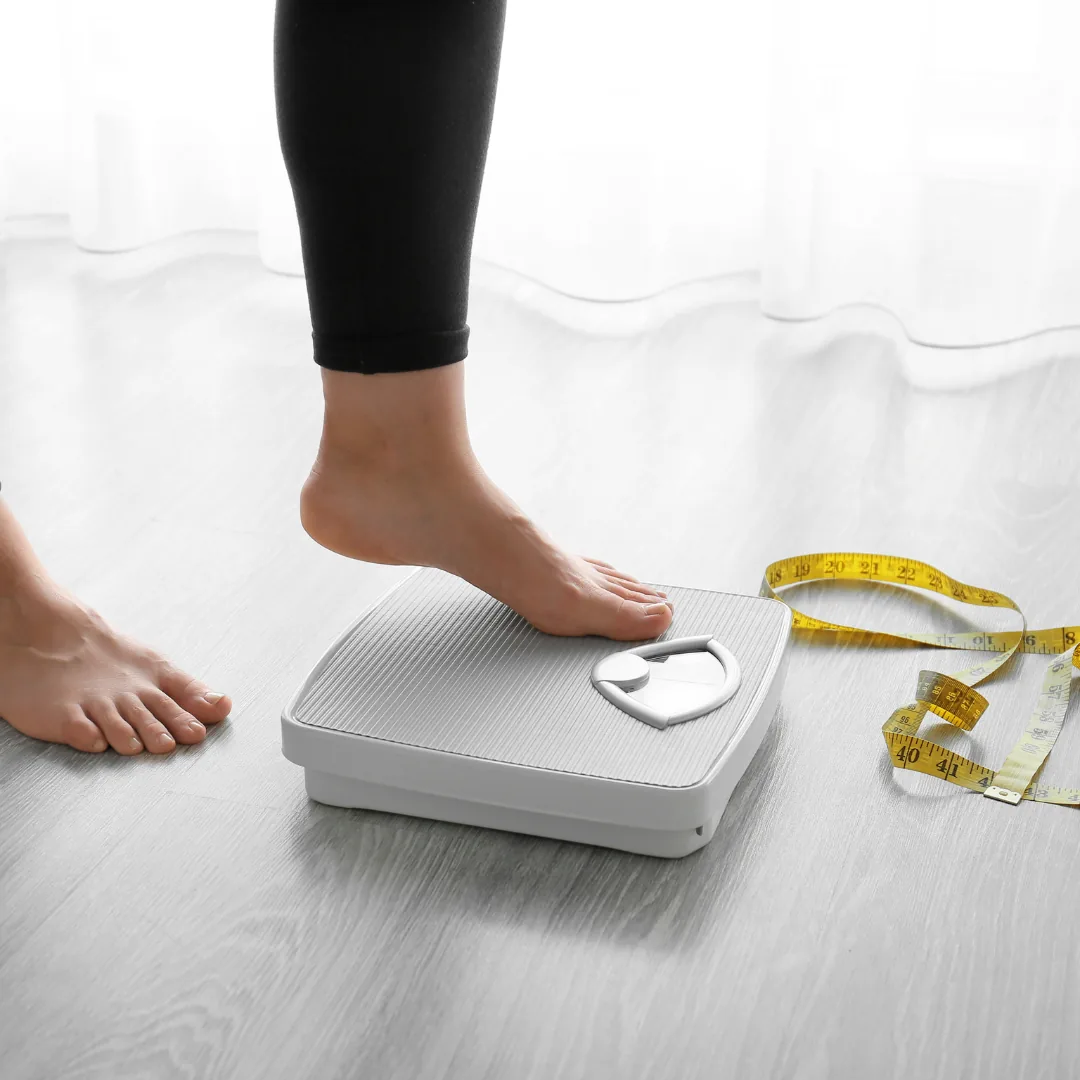 Person placing a foot on a bathroom scale beside a yellow measuring tape, illustrating weight monitoring often associated with thyroid health.