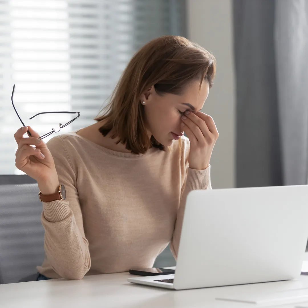 Woman rubbing her eyes and holding eyeglasses while seated at a laptop, appearing tired.