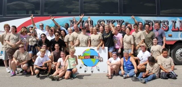 Large group of people posing in front of a bus, several holding a sign that reads "Love Has No Color".