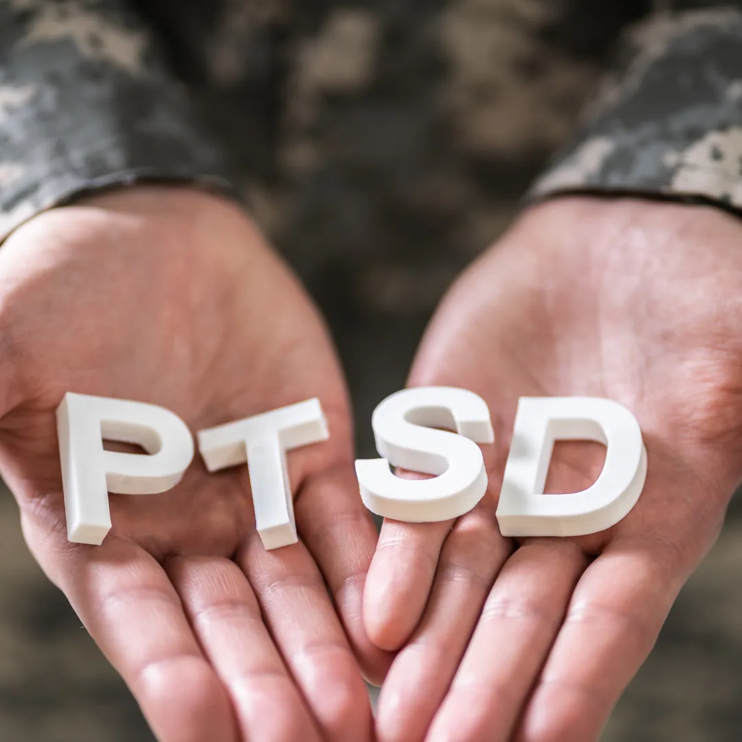 Open hands holding white block letters spelling PTSD, with camouflage clothing blurred in the background.