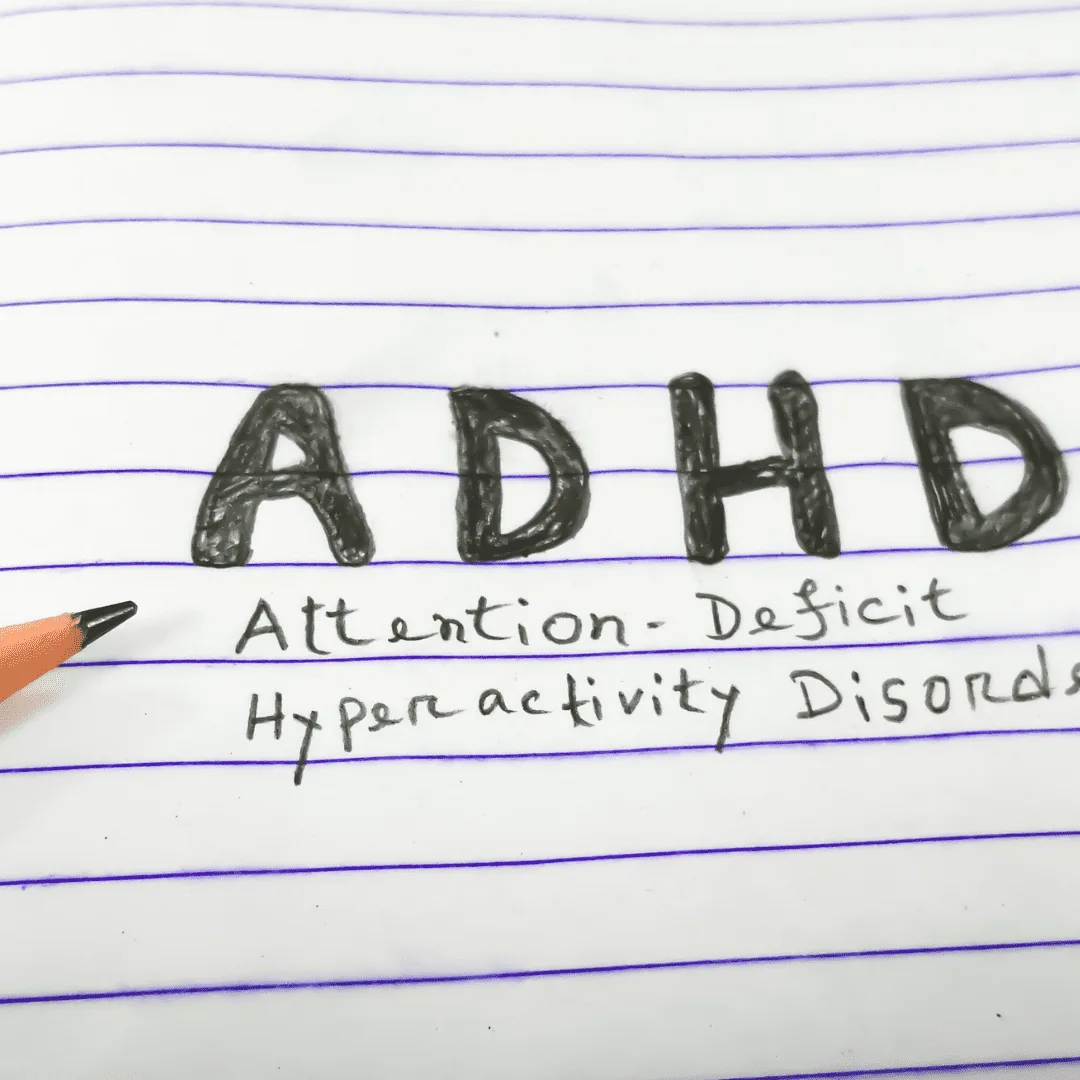 Handwritten 'ADHD' with the words 'Attention-Deficit Hyperactivity Disorder' on lined notebook paper, with a pencil tip pointing at the text.