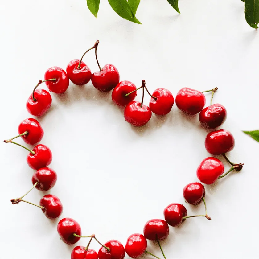 Cherries arranged in a heart shape on a white background with a few green leaves at the top.