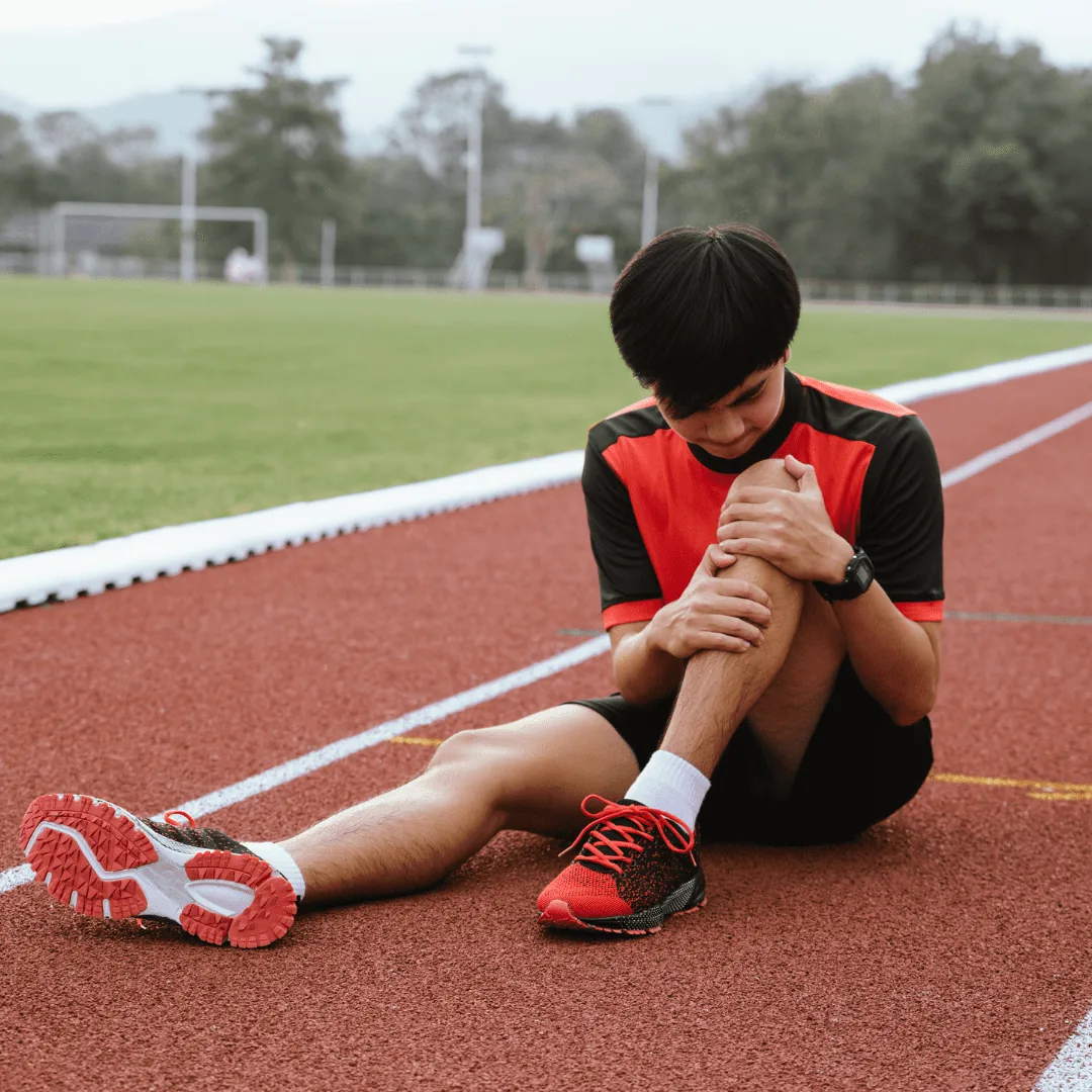 Athlete seated on a running track holding their knee while wearing a red training shirt and running shoes