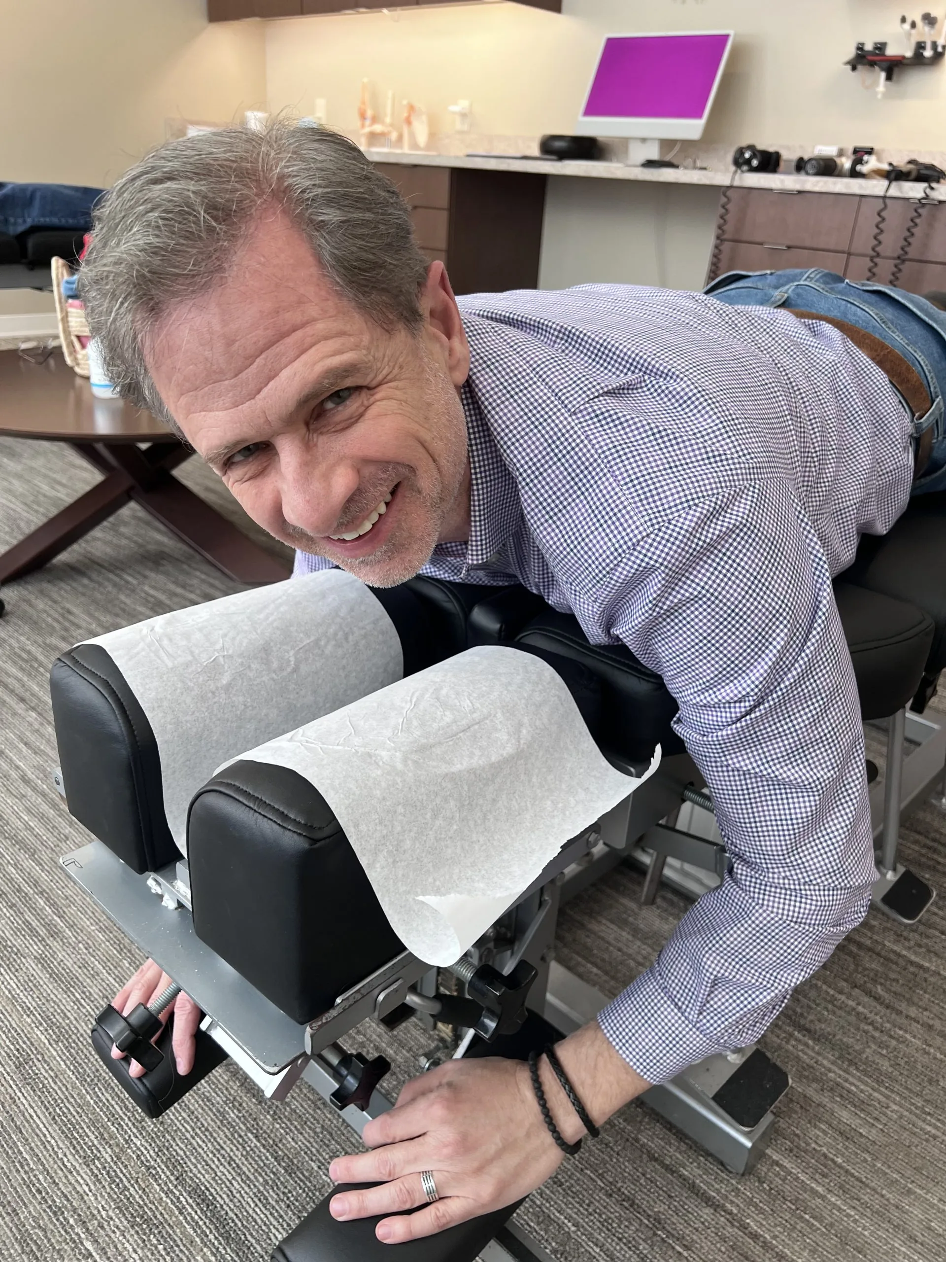 Man lying face down on a chiropractic adjustment table, smiling while reaching to adjust the table.