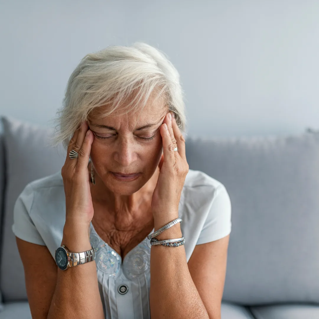 Older woman sitting on a couch with eyes closed, pressing her temples with both hands