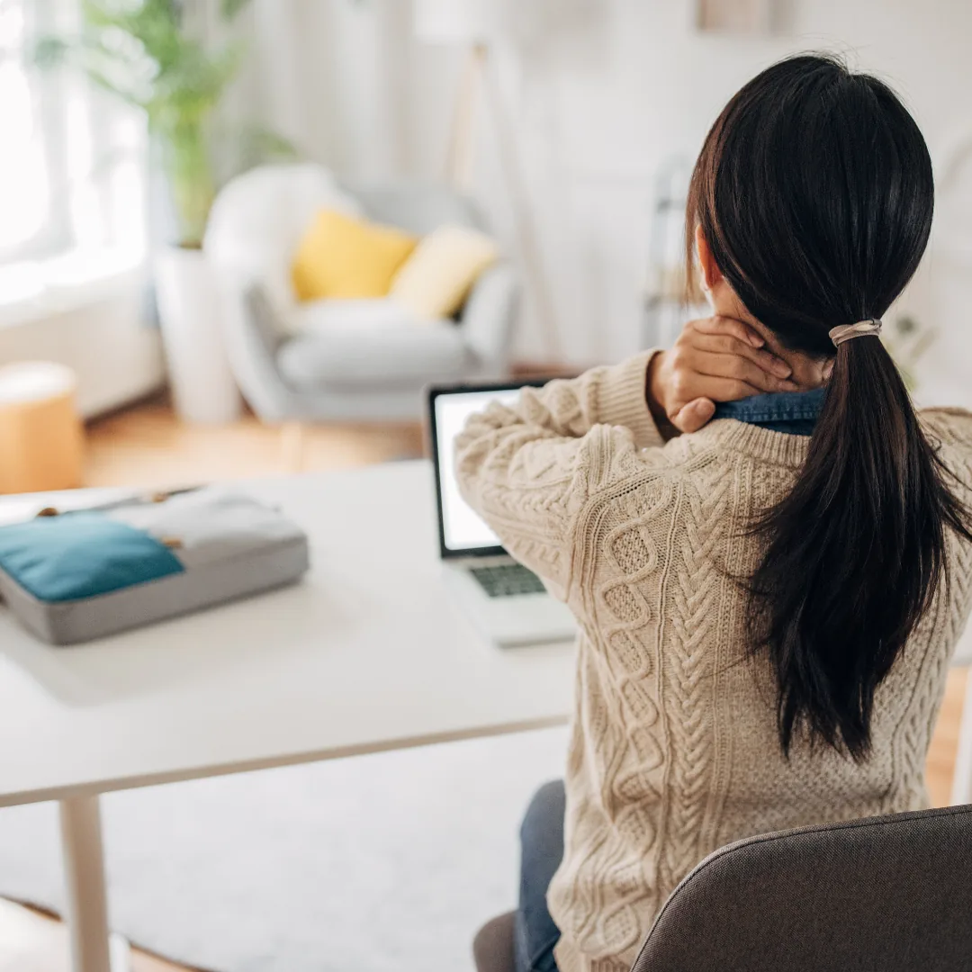 Person seated at a desk, facing away from the camera, holding their neck while working at a laptop in a home office.