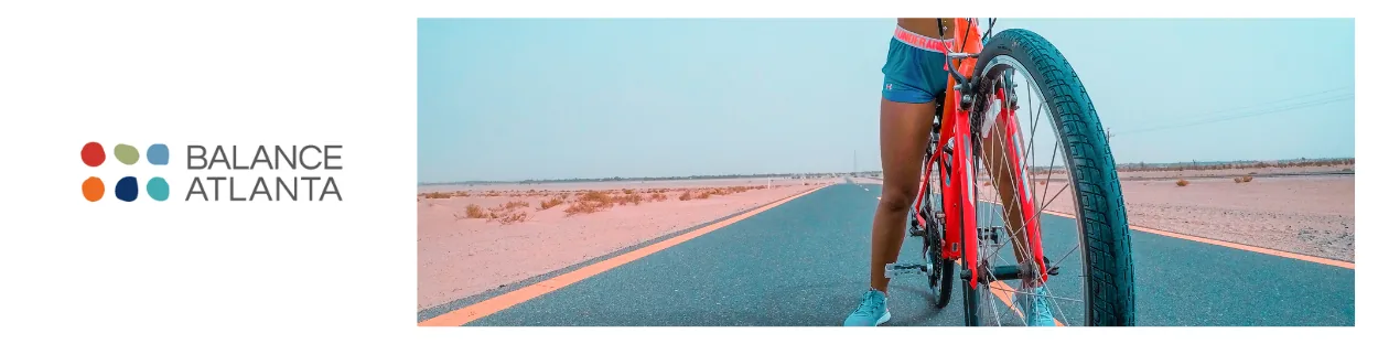 Legs of a person standing next to a red bicycle on a paved desert road with the Balance Atlanta logo at left.