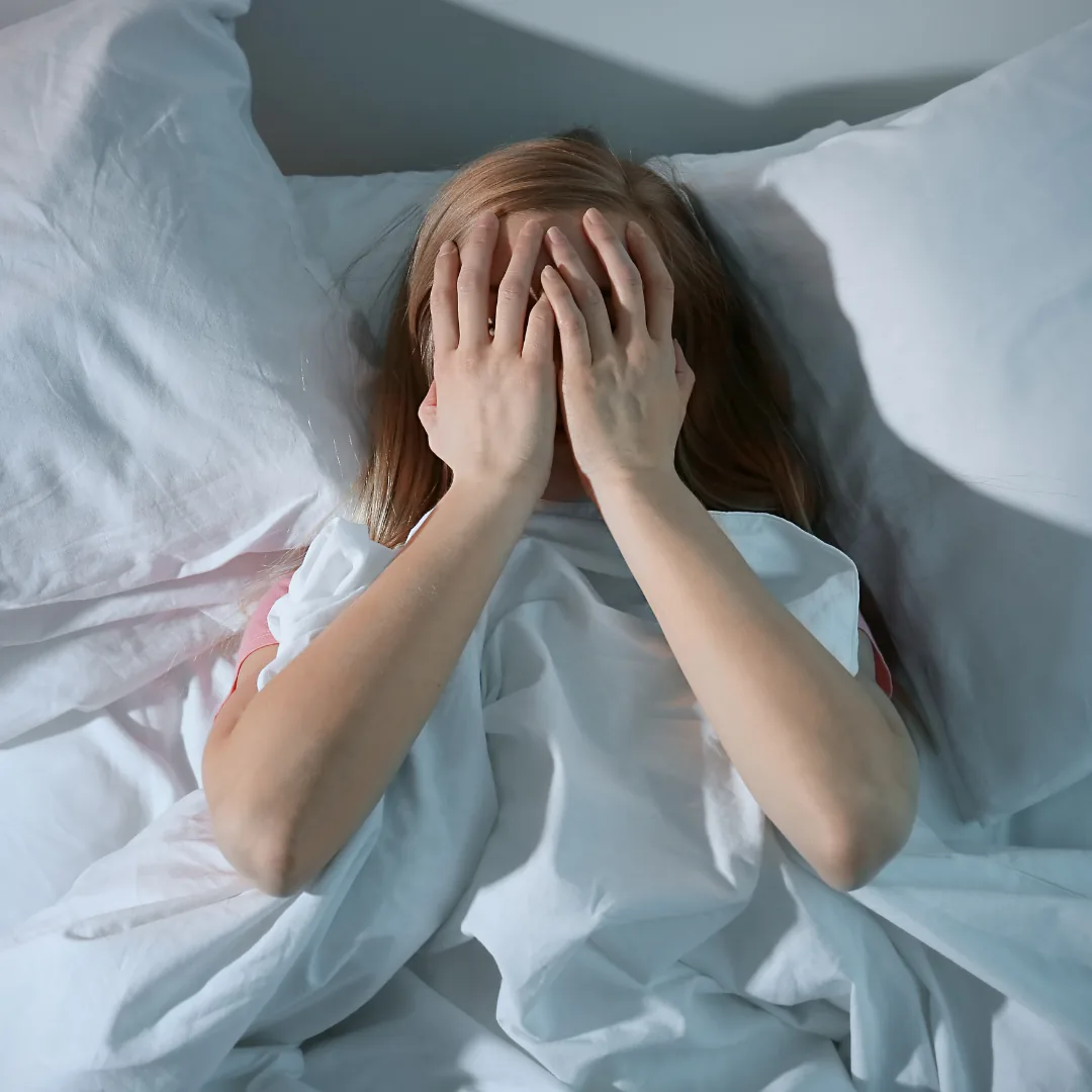 Person lying in bed under white sheets, covering their face with both hands.