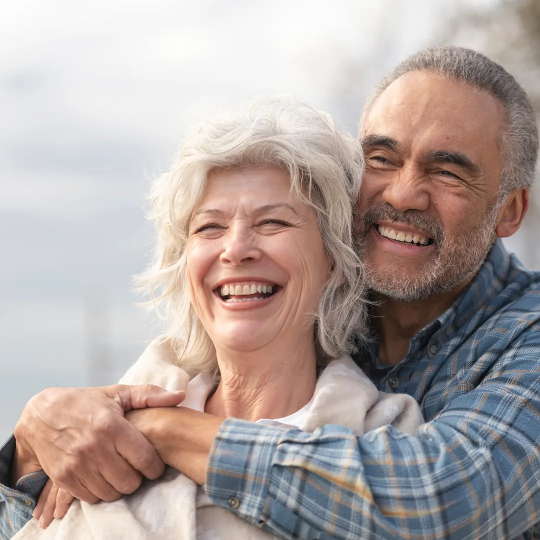 Smiling older couple outdoors with the man embracing the woman from behind as they laugh.