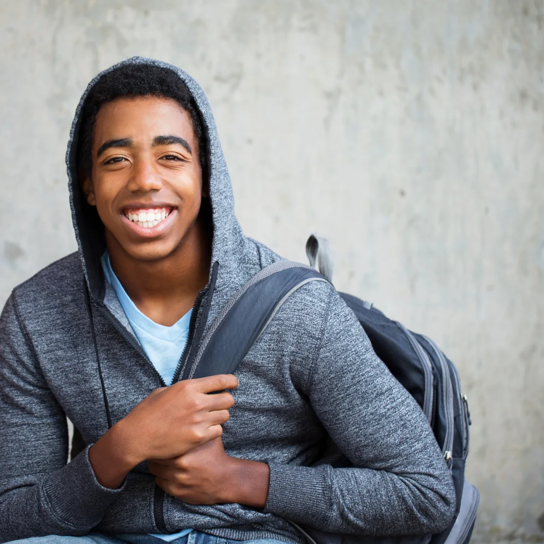 Smiling person in a gray hoodie holding a backpack strap while seated against a concrete wall.