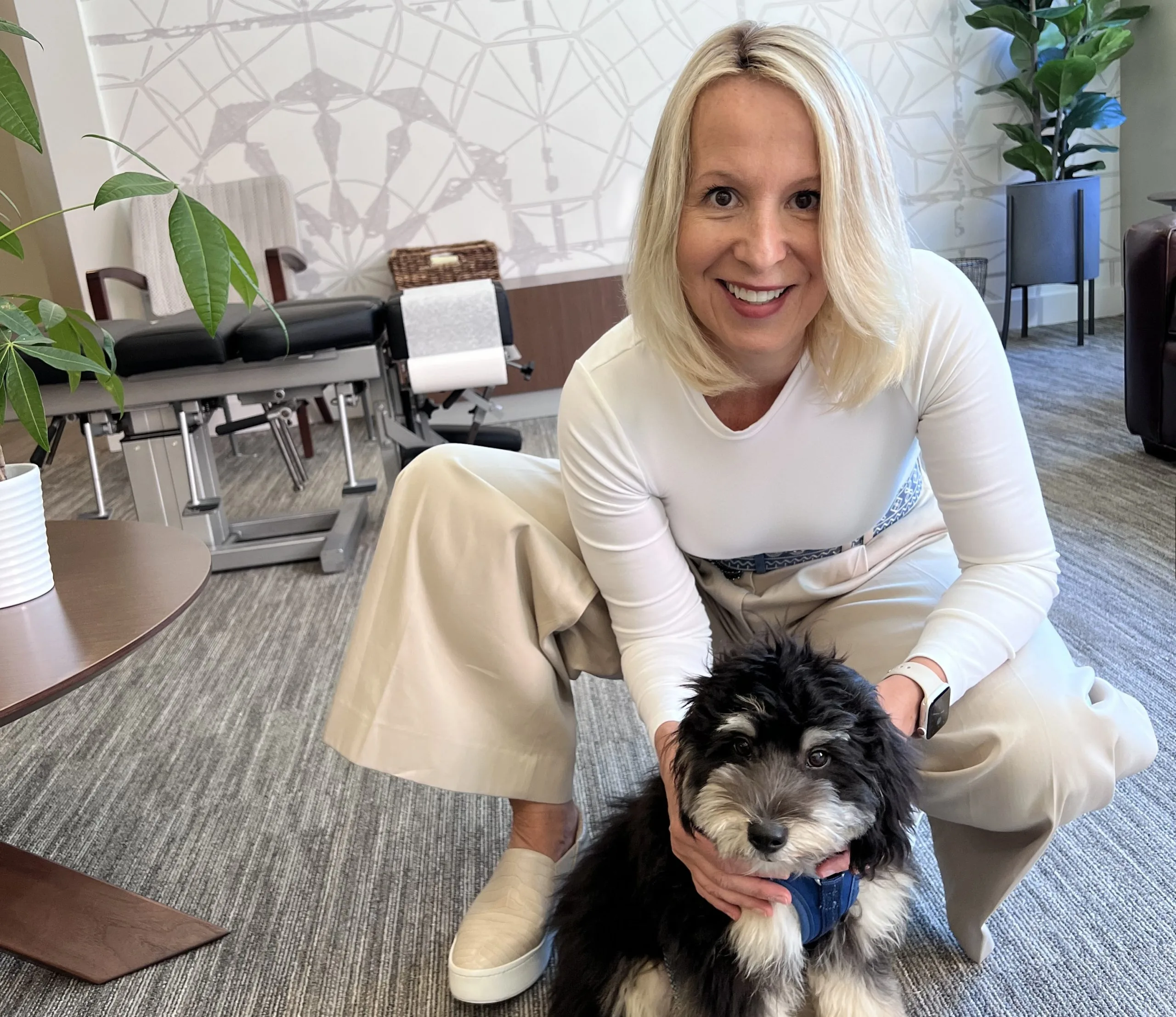 Smiling woman leaning toward camera while holding a small fluffy dog in a clinic treatment room.