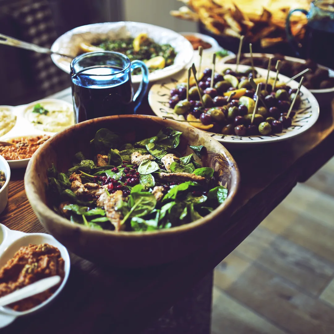wooden bowl of spinach salad with grilled chicken and pomegranate seeds on a table with bowls of olives and dips