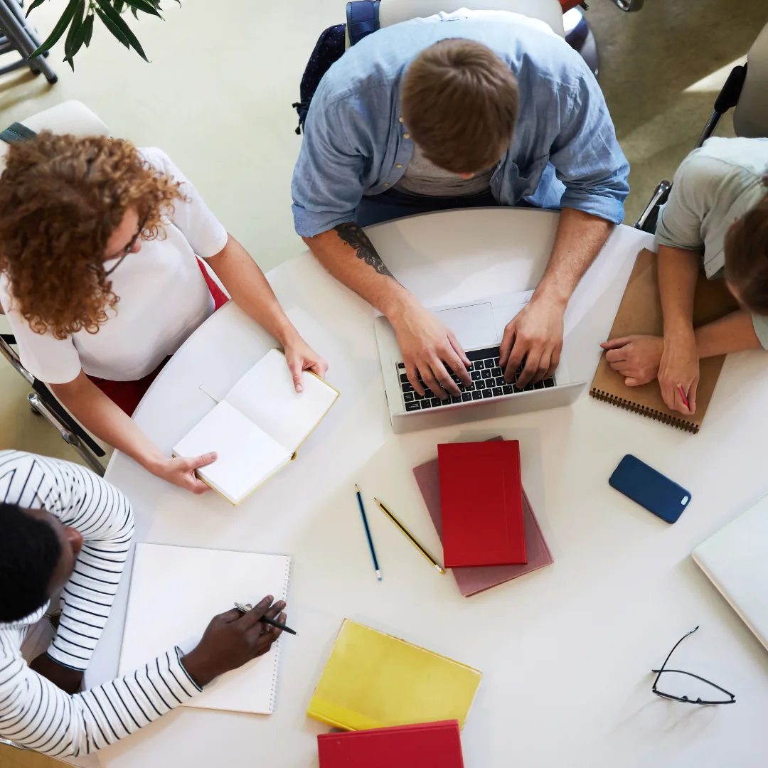 Overhead view of four people working around a round table with a laptop, notebooks, pencils, and a smartphone.