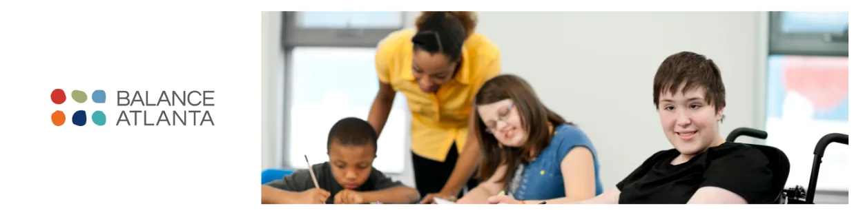 Teacher leaning over a table helping three students with schoolwork; a student at right smiles while seated in a wheelchair.