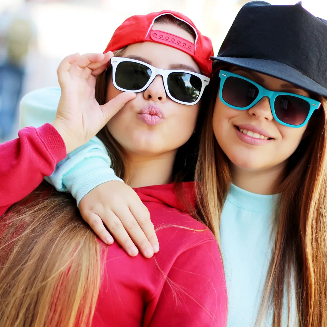 Two girls smiling and posing together while wearing sunglasses and baseball caps.