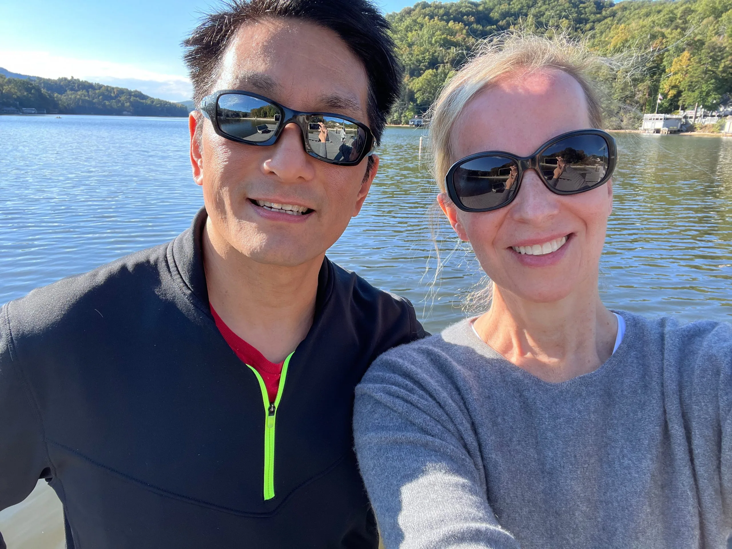 Two people wearing sunglasses smiling for a selfie on a lake with tree-covered hills in the background.