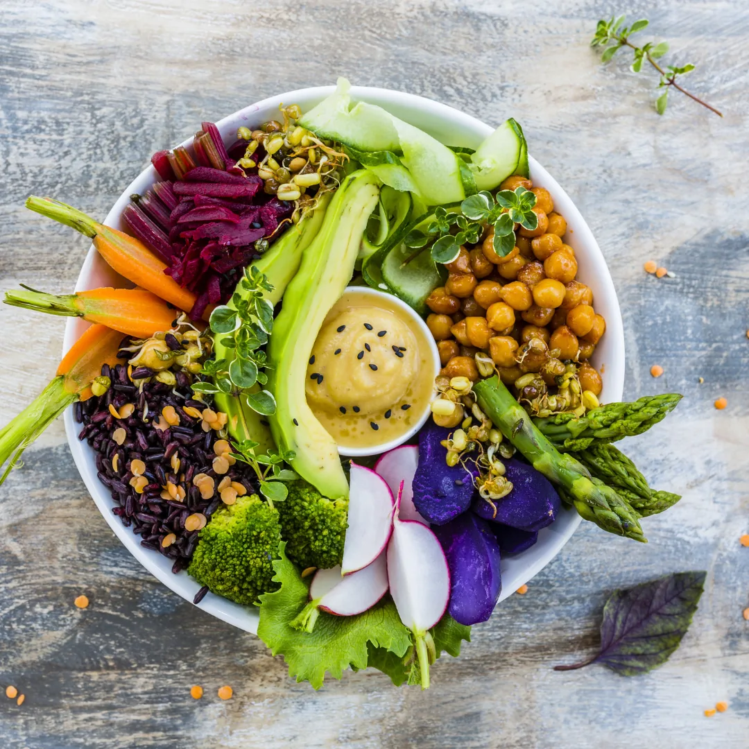 Bowl of mixed vegetables and grains with chickpeas, avocado, asparagus, radishes, purple potatoes, broccoli, carrots, black rice, sprouts, and a sesame-topped creamy dressing.