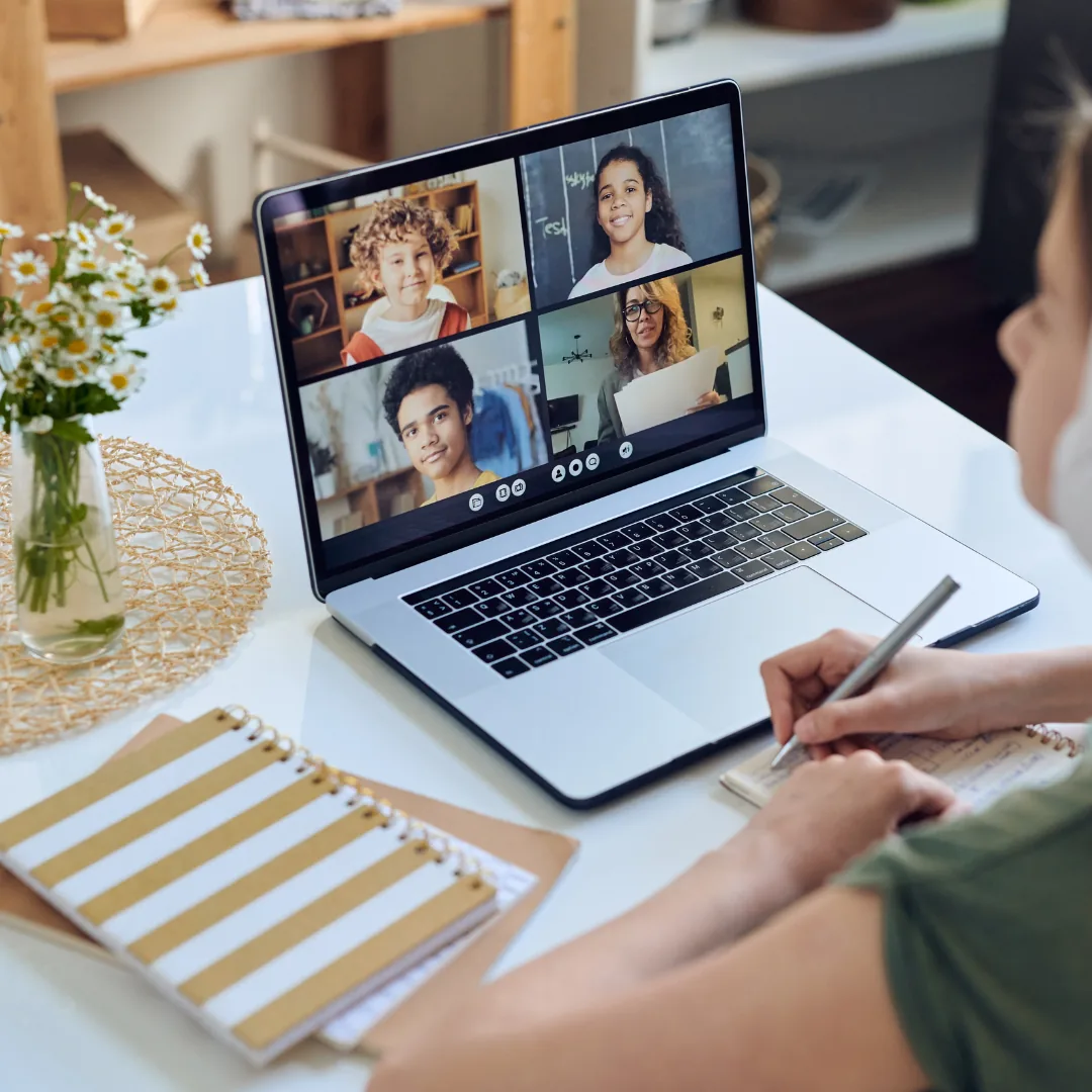 Laptop screen displaying a four-person video call while someone at a desk writes notes.