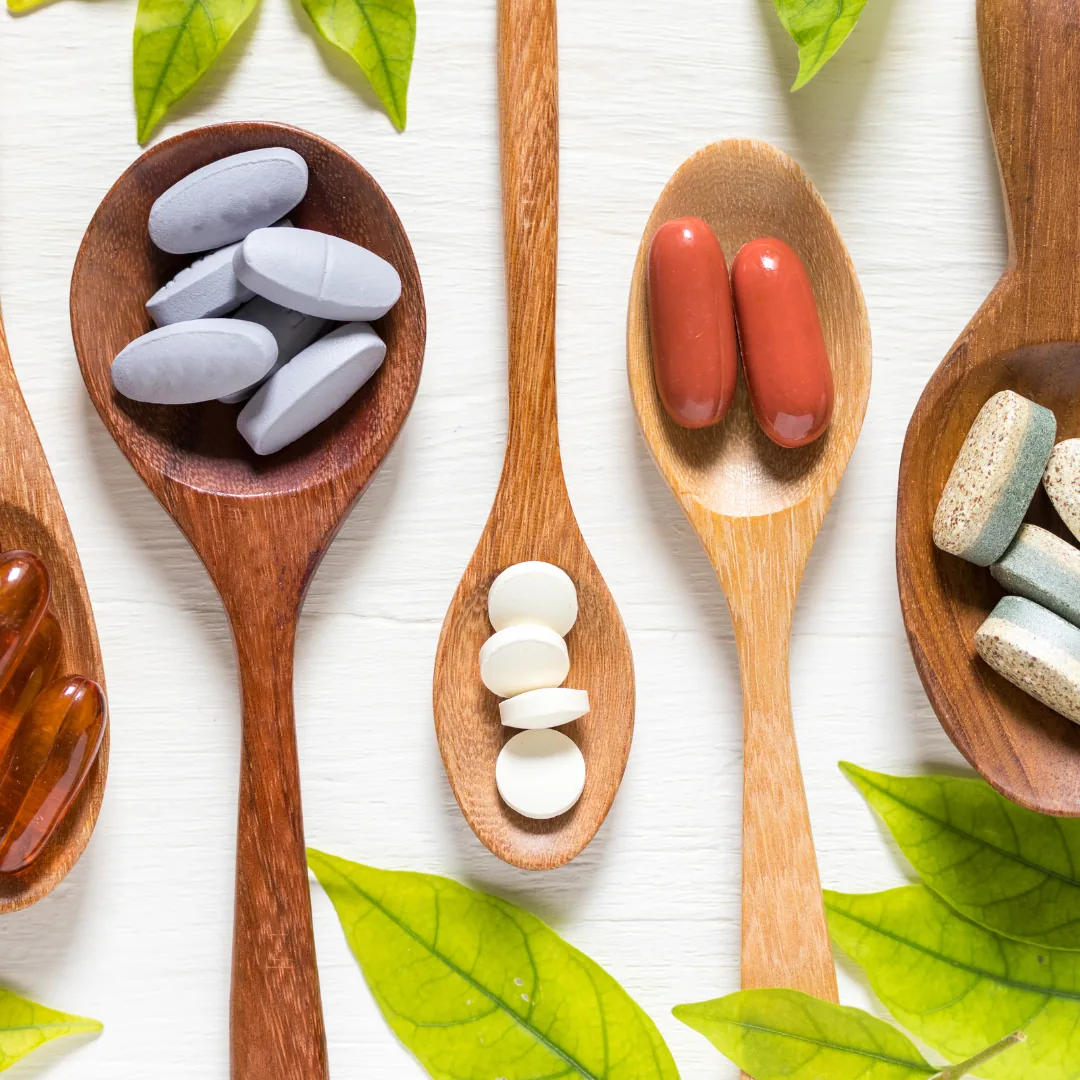 Wooden spoons holding various vitamin and mineral supplement tablets and capsules arranged on a white surface with green leaves.