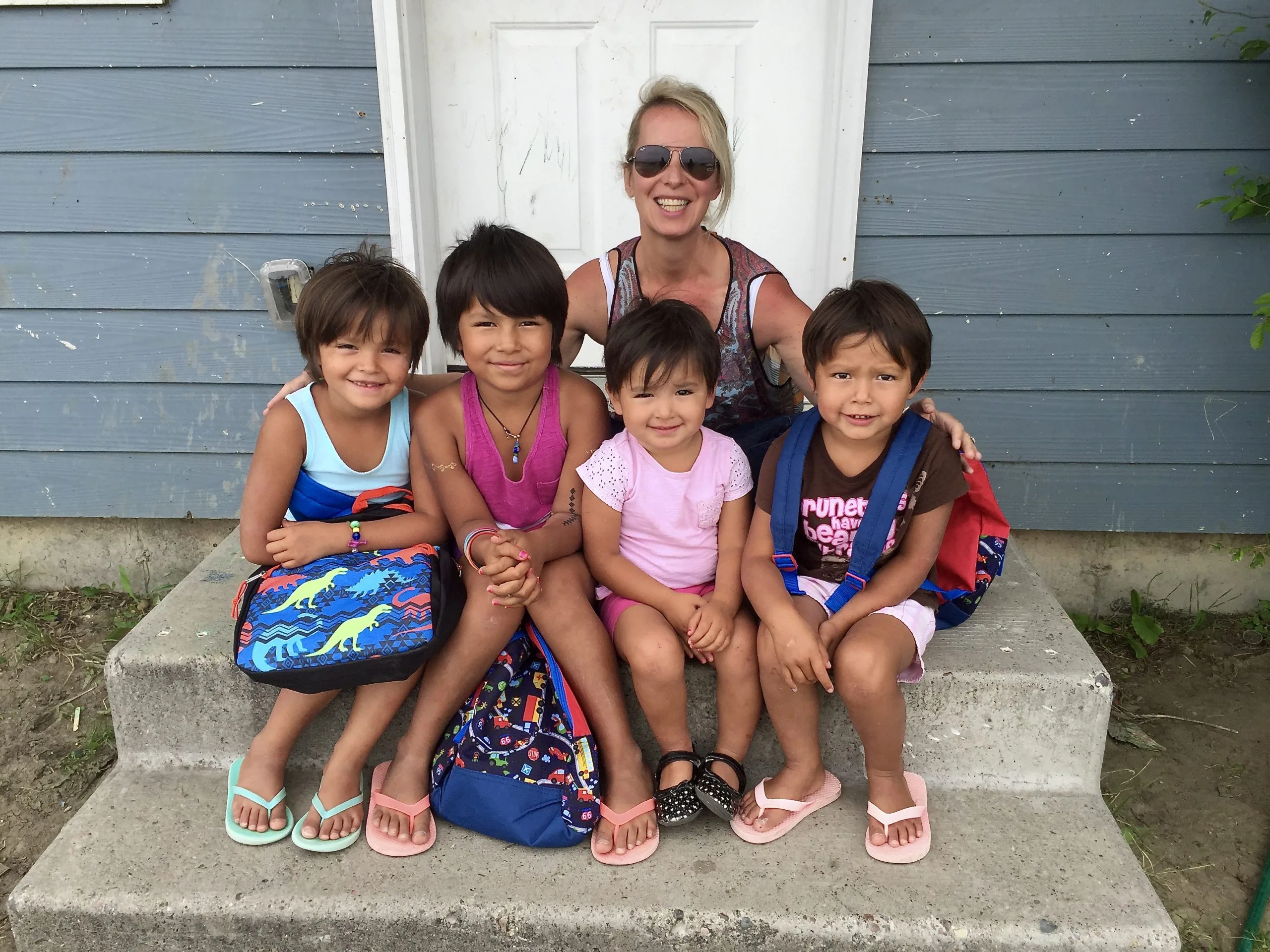 Smiling woman kneeling behind four children seated on concrete steps with backpacks.