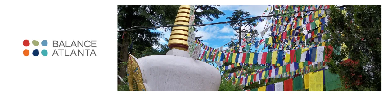 White Buddhist stupa with a golden spire beside multicolored Tibetan prayer flags strung among trees under a blue sky.