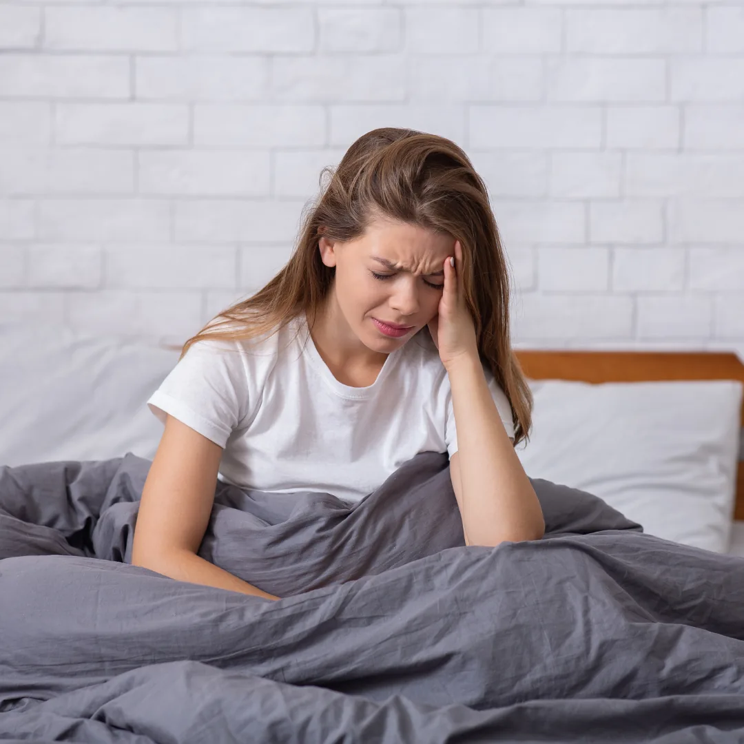 Woman sitting up in bed with a pained expression, holding her head and appearing tired or distressed.