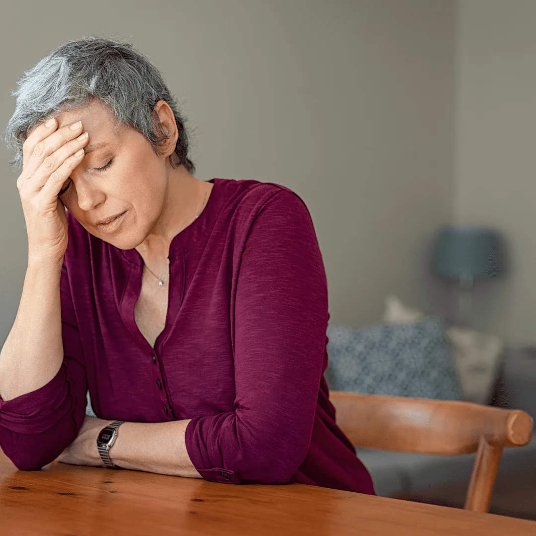 Woman sitting at a table with eyes closed, holding her forehead and resting an arm on the table.