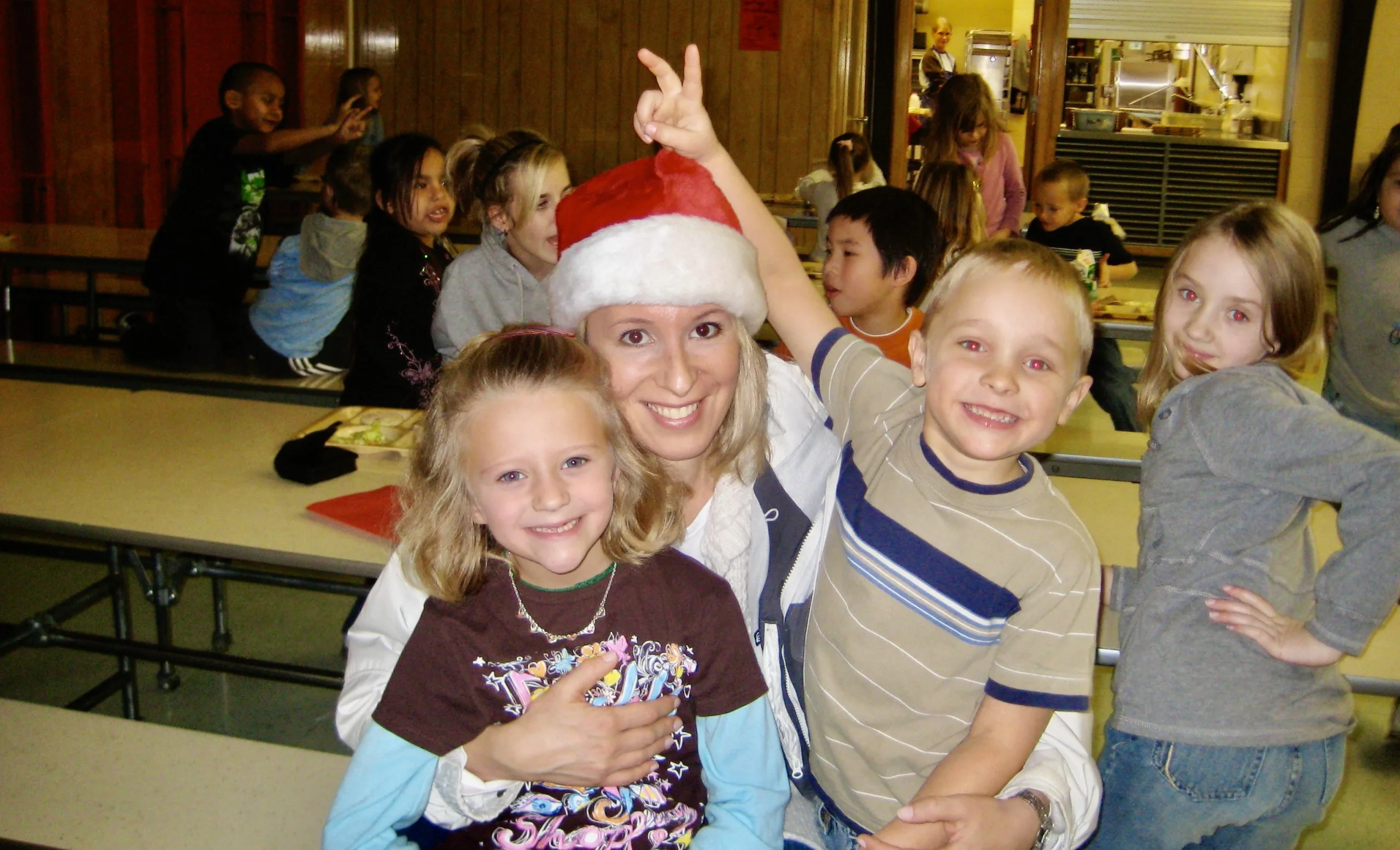Adult wearing a Santa hat smiling and posing with several children in a school cafeteria during a holiday event.