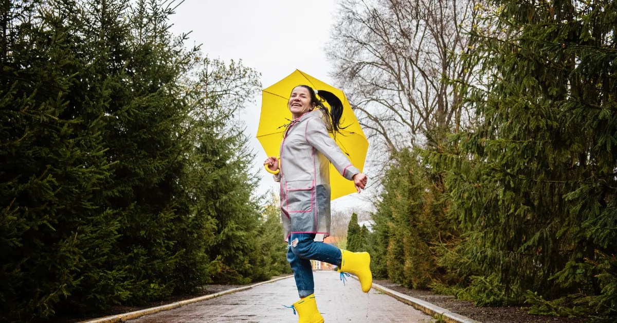 Woman jumping on a wet park path between evergreen trees, smiling and holding a yellow umbrella while wearing yellow rain boots and a gray raincoat.