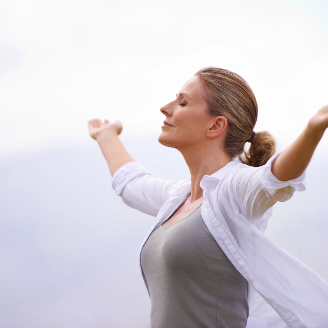 Woman standing outdoors with arms outstretched and eyes closed, looking relaxed.